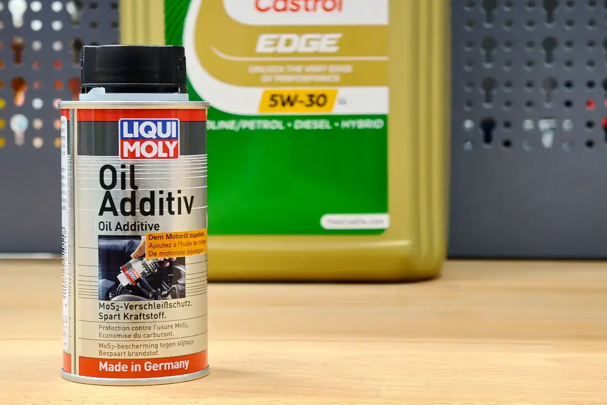Motoröl Additive: Eine silberne Metalldose des Zusatzmittels "Liqui Moly Oil Additiv" steht im Vordergrund auf einer hellen Arbeitsfläche. Im unscharfen Hintergrund ist eine große, goldfarbene Plastikflasche von "Castrol Edge 5W-30" Motoröl sowie eine dunkelgraue Lochwand zu sehen. Das Licht ist hell und gleichmäßig, die Atmosphäre gleicht einer Werkstatt.
