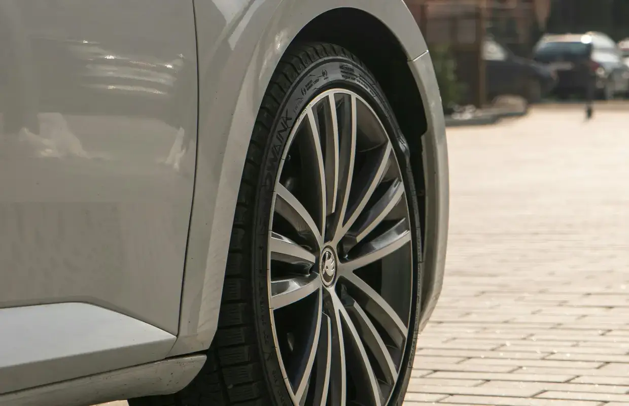 Skoda Reifengrößen. Nahaufnahme des vorderen linken Rades eines silbernen oder lichtgrauen Skoda-Fahrzeugs auf gepflastertem Untergrund. Zu sehen ist eine filigrane Vielspeichen-Leichtmetallfelge in Bi-Color-Optik (Silber/Schwarz) mit dem Skoda-Logo auf der Nabenkappe. Der montierte Reifen zeigt ein asymmetrisches Profil und Markierungen auf der Flanke. Das natürliche Tageslicht fällt von rechts ein und erzeugt weiche Schatten im Radkasten sowie Reflexionen auf dem Lack.